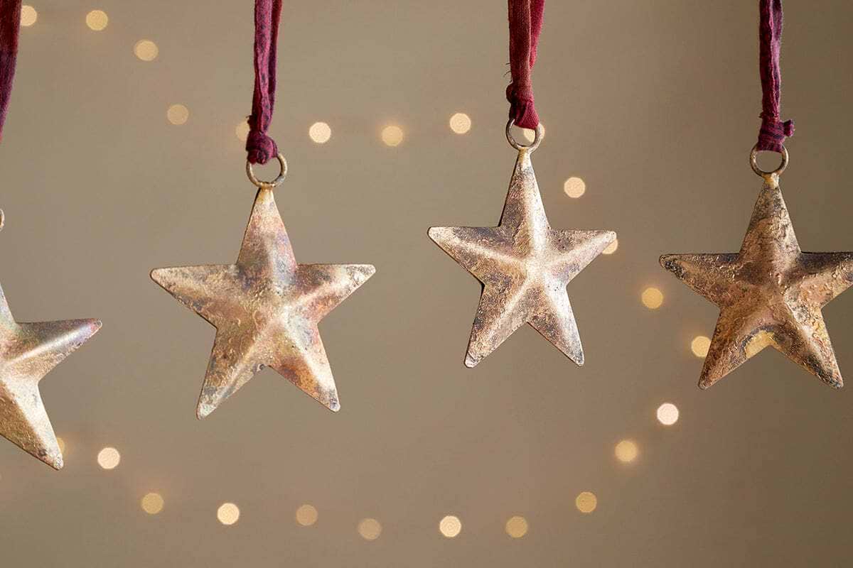 The nkuku Roshani Star Baubles - Antique Brass (Set of 4) hang from red strings against a beige backdrop with soft string lights. Only parts of the two outer star silhouettes are visible at the edges, creating a warm, festive feel.