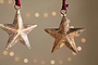 The Roshani Star Baubles - Antique Brass (Set of 4) by nkuku feature rustic, gold-toned stars with a textured, weathered finish. Hanging from red strings, they create a festive holiday feel against soft beige backgrounds.