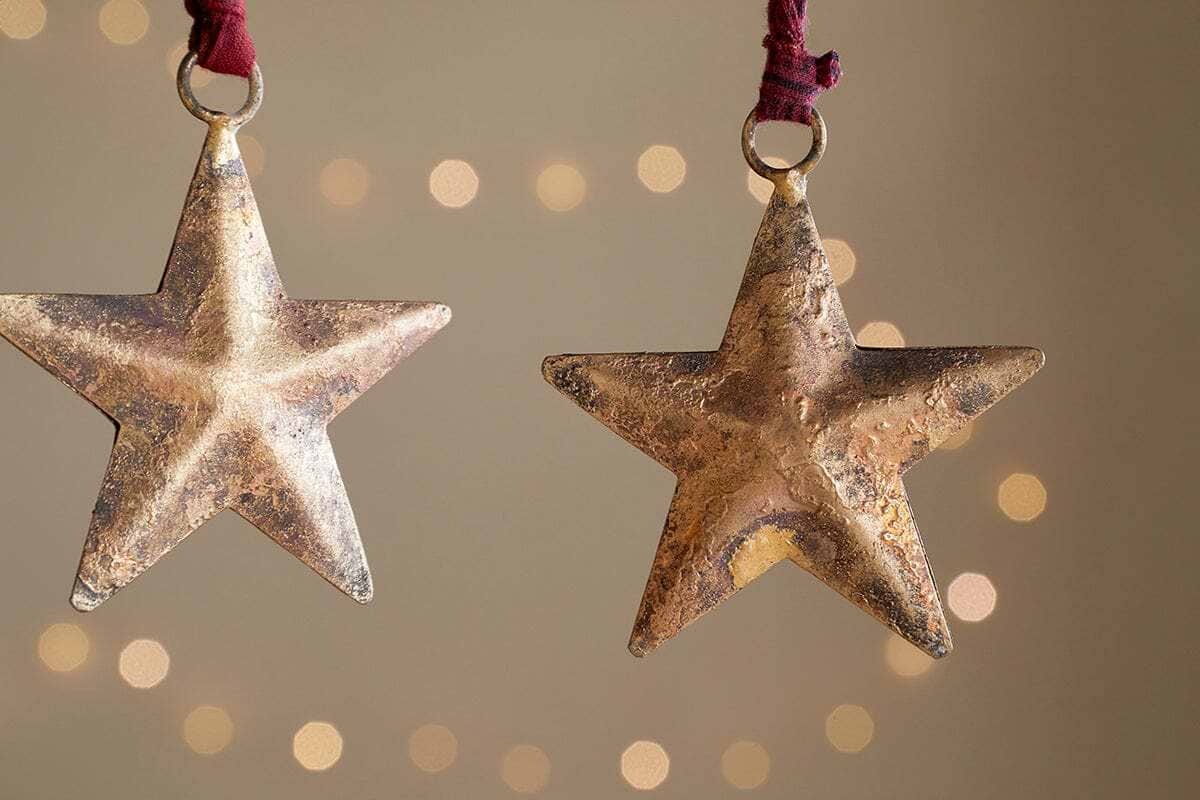 The Roshani Star Baubles - Antique Brass (Set of 4) by nkuku feature rustic, gold-toned stars with a textured, weathered finish. Hanging from red strings, they create a festive holiday feel against soft beige backgrounds.