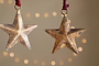 The Roshani Star Baubles - Antique Brass (Set of 4) by nkuku feature rustic, gold-toned stars with a textured, weathered finish. Hanging from red strings, they create a festive holiday feel against soft beige backgrounds.