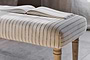 An open book rests on the nkuku Sanja Stripe Upholstered Bench in Grey, featuring natural cotton upholstery with slim dark stripes and wooden legs, placed at the end of a bed on a neutral-toned floor in a softly lit room.
