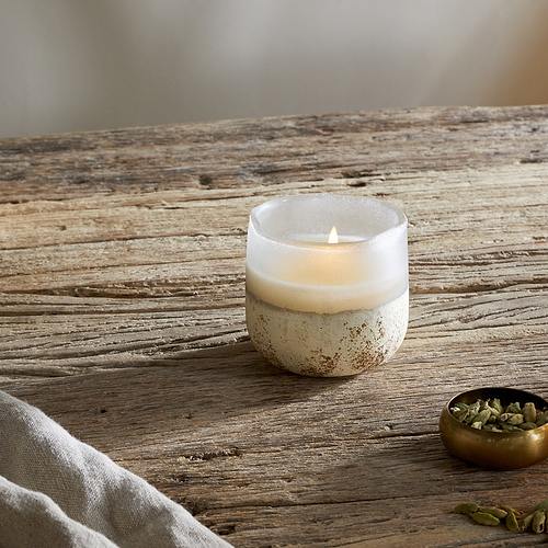A lit nkuku Cardamom & Amber 1 Wick Scented Candle in a frosted glass holder sits on a rustic wooden table beside a brass bowl of green cardamom pods, some loose pods, a cloth, and the candle’s labeled box.