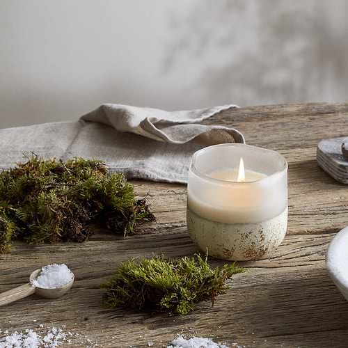 A nkuku Sea Salt & Moss 1 Wick Scented Candle in a frosted holder rests on a rustic wooden table with moss and a beige cloth, adding bespoke fragrance for a natural, calming atmosphere.