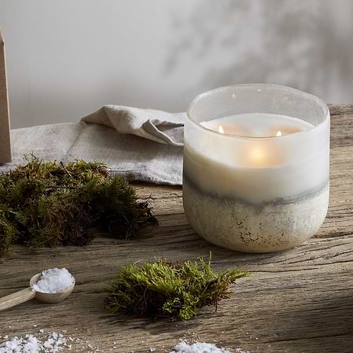 The nkuku Sea Salt & Moss 3 Wick Scented Candle, in a white and gray marbled holder, rests on a rustic wooden table beside green moss, sea salt, and its box—evoking a bespoke UK-inspired fragrance.