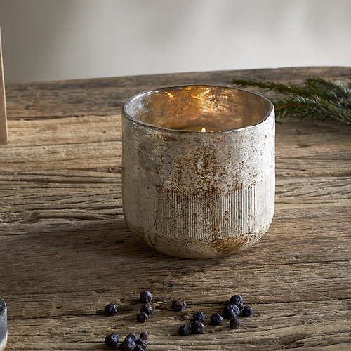 The nkuku Juniper & Fir 3 Wick Scented Candle in a rustic glass holder sits on a wooden table with juniper berries, a pinecone, and fir branch—a cozy display that captures the essence of winter.