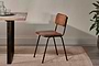 The Ukari Leather Dining Chair - Aged Tan by nkuku, featuring a padded leather seat and metal frame, sits beside a wooden table on a beige woven rug. Neutral tones and a visible plant add to the space's modern, utilitarian style.