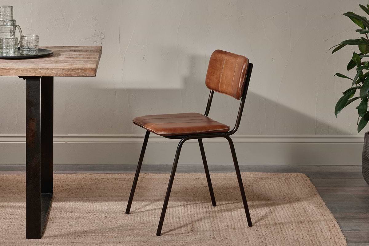 The Ukari Leather Dining Chair - Aged Tan by nkuku, featuring a padded leather seat and metal frame, sits beside a wooden table on a beige woven rug. Neutral tones and a visible plant add to the space's modern, utilitarian style.