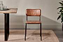 The Ukari Leather Dining Chair - Aged Tan by nkuku, featuring a padded seat, square backrest, and slim black metal legs, sits on a beige rug beside a wooden table with glassware and a potted plant against a light wall.