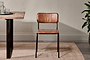 The Ukari Leather Dining Chair - Aged Tan by nkuku, featuring a padded seat, square backrest, and slim black metal legs, sits on a beige rug beside a wooden table with glassware and a potted plant against a light wall.