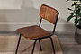 The nkuku Ukari Leather Dining Chair in Aged Tan features a padded leather seat, visible stitching, and black metal legs. It sits on wooden flooring near a woven rug, with a light wall and a plant in a gray pot in the background.