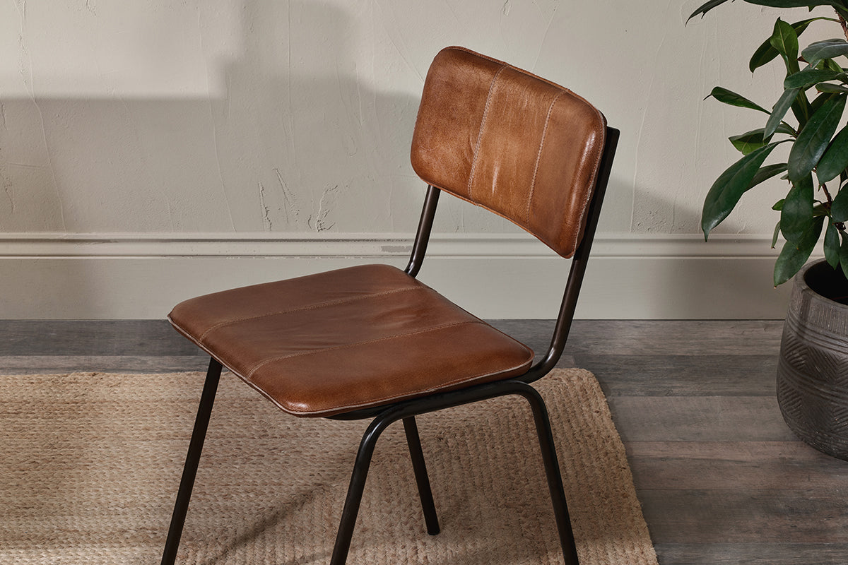 The nkuku Ukari Leather Dining Chair in Aged Tan features a padded leather seat, visible stitching, and black metal legs. It sits on wooden flooring near a woven rug, with a light wall and a plant in a gray pot in the background.
