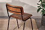 The Ukari Leather Dining Chair - Aged Tan from nkuku, featuring a padded brown leather seat and black metal frame, stands on a tan woven rug atop a wooden floor. A leafy green potted plant sits by the cream-colored textured wall.