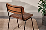 The Ukari Leather Dining Chair - Aged Tan from nkuku, featuring a padded brown leather seat and black metal frame, stands on a tan woven rug atop a wooden floor. A leafy green potted plant sits by the cream-colored textured wall.