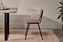 The nkuku Ukari Leather Dining Chair - Aged Tan, with its padded leather seat and black metal frame, sits by a rustic wooden table on a woven beige rug. Clear glasses top the table, with a textured off-white wall and green plant nearby.