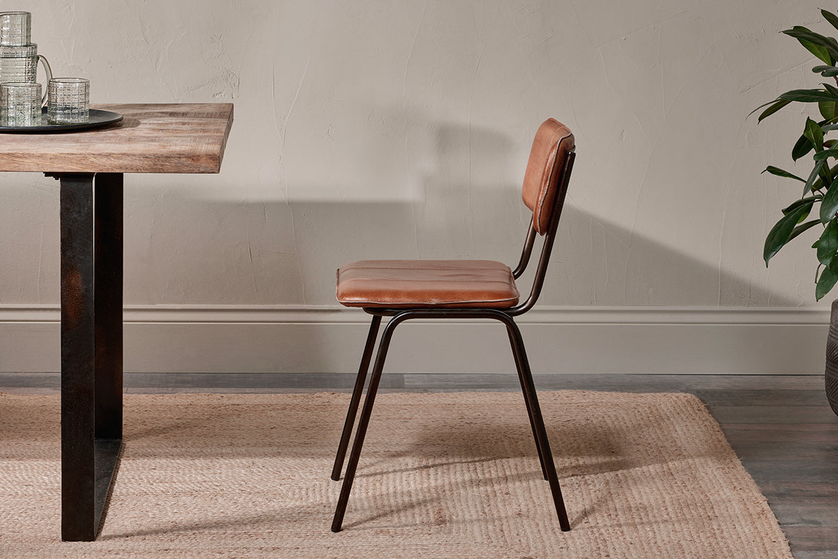 The nkuku Ukari Leather Dining Chair - Aged Tan, with its padded leather seat and black metal frame, sits by a rustic wooden table on a woven beige rug. Clear glasses top the table, with a textured off-white wall and green plant nearby.