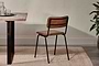 The Ukari Leather Dining Chair by nkuku in aged tan leather, featuring a padded seat and black metal frame, sits beside a rustic wooden table with clear glasses on a dark tray, atop a beige rug, with a potted plant and neutral wall.