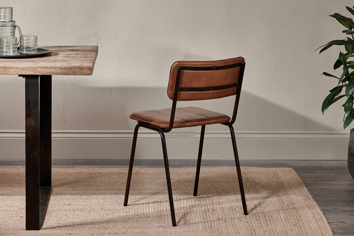 The Ukari Leather Dining Chair by nkuku in aged tan leather, featuring a padded seat and black metal frame, sits beside a rustic wooden table with clear glasses on a dark tray, atop a beige rug, with a potted plant and neutral wall.