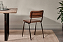 The Ukari Leather Dining Chair by nkuku in aged tan leather, featuring a padded seat and black metal frame, sits beside a rustic wooden table with clear glasses on a dark tray, atop a beige rug, with a potted plant and neutral wall.