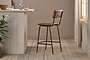 The Ukari Counter Chair - Aged Tan by nkuku, featuring a metal frame, stands on light wooden floors by a rustic pale wood counter topped with glass tumblers, a decanter, and small plants. Sunlight filters through sheer curtains from the right.