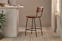 The Ukari Counter Chair - Aged Tan by nkuku, featuring a metal frame, stands on light wooden floors by a rustic pale wood counter topped with glass tumblers, a decanter, and small plants. Sunlight filters through sheer curtains from the right.