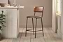 The nkuku Ukari Counter Chair in Aged Tan leather with a metal frame stands on a light wood floor by a rustic counter. Sunlight filters through sheer curtains, highlighting green plants and glassware arranged near the window.