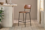 The nkuku Ukari Counter Chair in Aged Tan leather with a metal frame stands on a light wood floor by a rustic counter. Sunlight filters through sheer curtains, highlighting green plants and glassware arranged near the window.