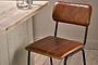 The Ukari Counter Chair - Aged Tan by nkuku, featuring visible stitching and black metal legs, sits beside a light wood table topped with two glass cups, all set on pale wooden plank flooring.