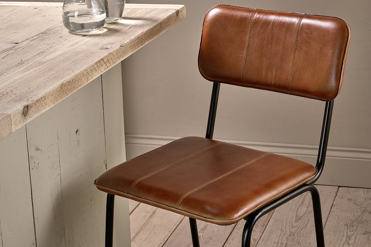 The Ukari Counter Chair - Aged Tan by nkuku, featuring visible stitching and black metal legs, sits beside a light wood table topped with two glass cups, all set on pale wooden plank flooring.