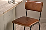 The Ukari Counter Chair - Aged Tan by nkuku, featuring visible stitching and black metal legs, sits beside a light wood table topped with two glass cups, all set on pale wooden plank flooring.