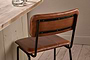 The Ukari Counter Chair in aged tan leather from nkuku, featuring black metal legs and backrest supports, sits by a rustic wooden table set with two glasses of water. The scene includes light wood flooring and a beige wall with simple base molding.