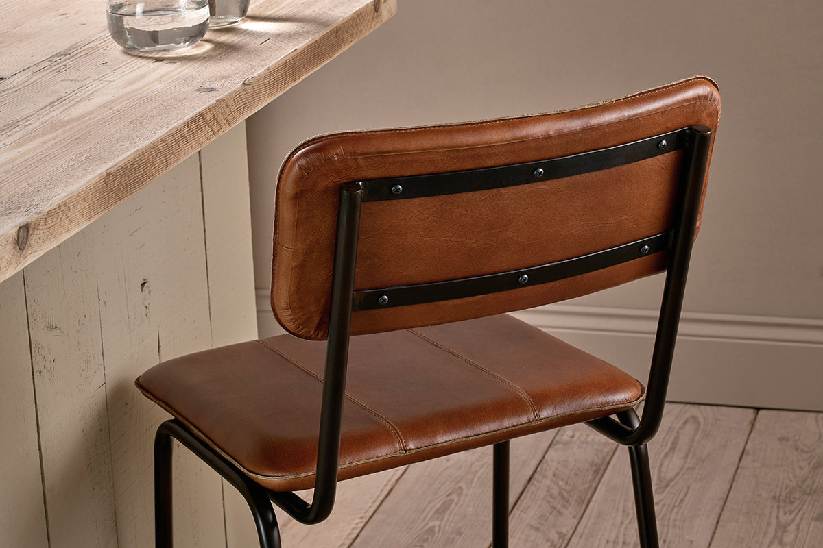 The Ukari Counter Chair in aged tan leather from nkuku, featuring black metal legs and backrest supports, sits by a rustic wooden table set with two glasses of water. The scene includes light wood flooring and a beige wall with simple base molding.