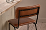 The Ukari Counter Chair in aged tan leather from nkuku, featuring black metal legs and backrest supports, sits by a rustic wooden table set with two glasses of water. The scene includes light wood flooring and a beige wall with simple base molding.