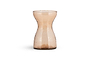 The nkuku Enir Hyacinth Vase in Dusty Terracotta features a tall, narrow, hourglass design with lightly hammered glass for a hand-blown look. Displayed on a white background, it casts a subtle shadow beneath it.