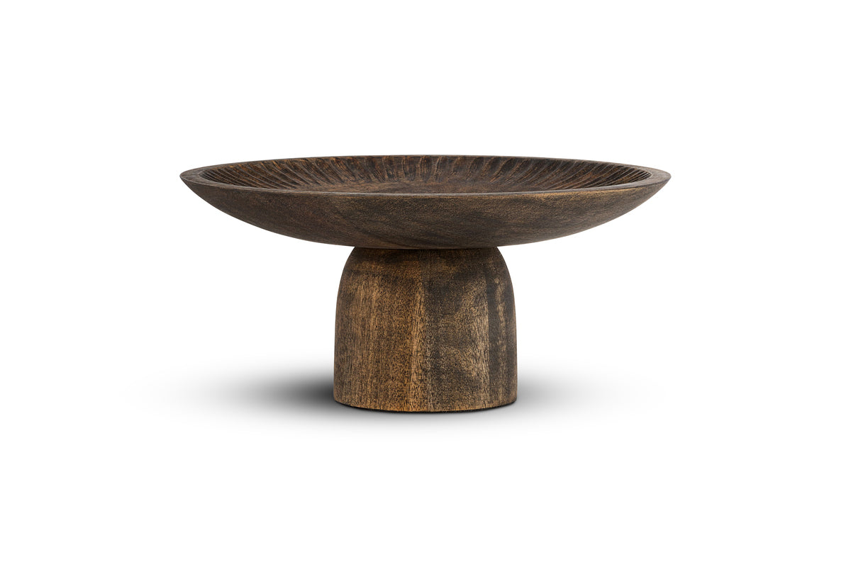 The nkuku Eisha Wooden Cake Stand in brown features a hand-carved, wide shallow dish on a sturdy cylindrical base. Its textured surface and natural grain patterns make it ideal for serving or as a rustic cake stand. Photographed on white background.