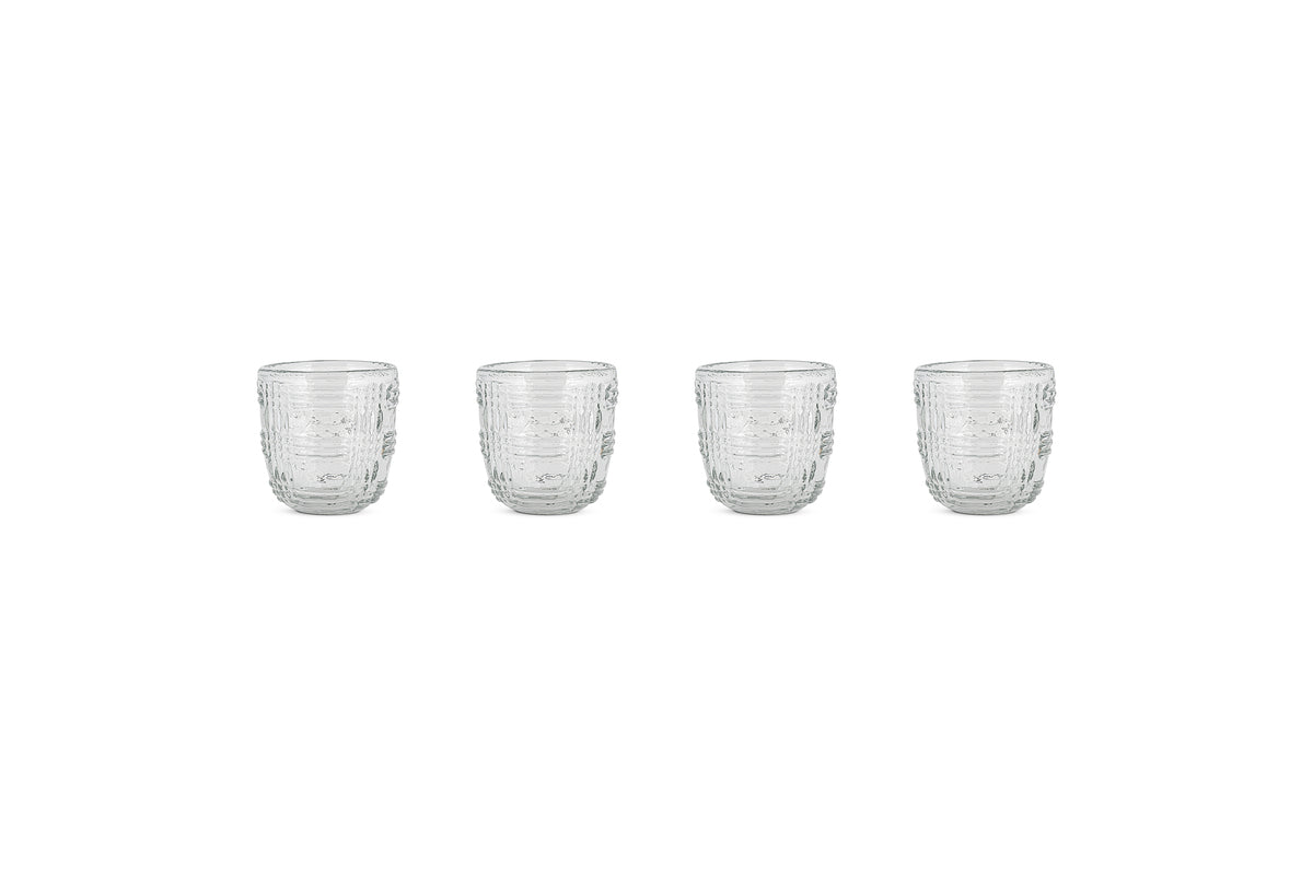 Four nkuku Kaanch Tumblers (Set of 4, Clear) are arranged in a horizontal row on a white background, each empty and evenly spaced. The glass tumblers feature an embossed check pattern with a textured geometric design.