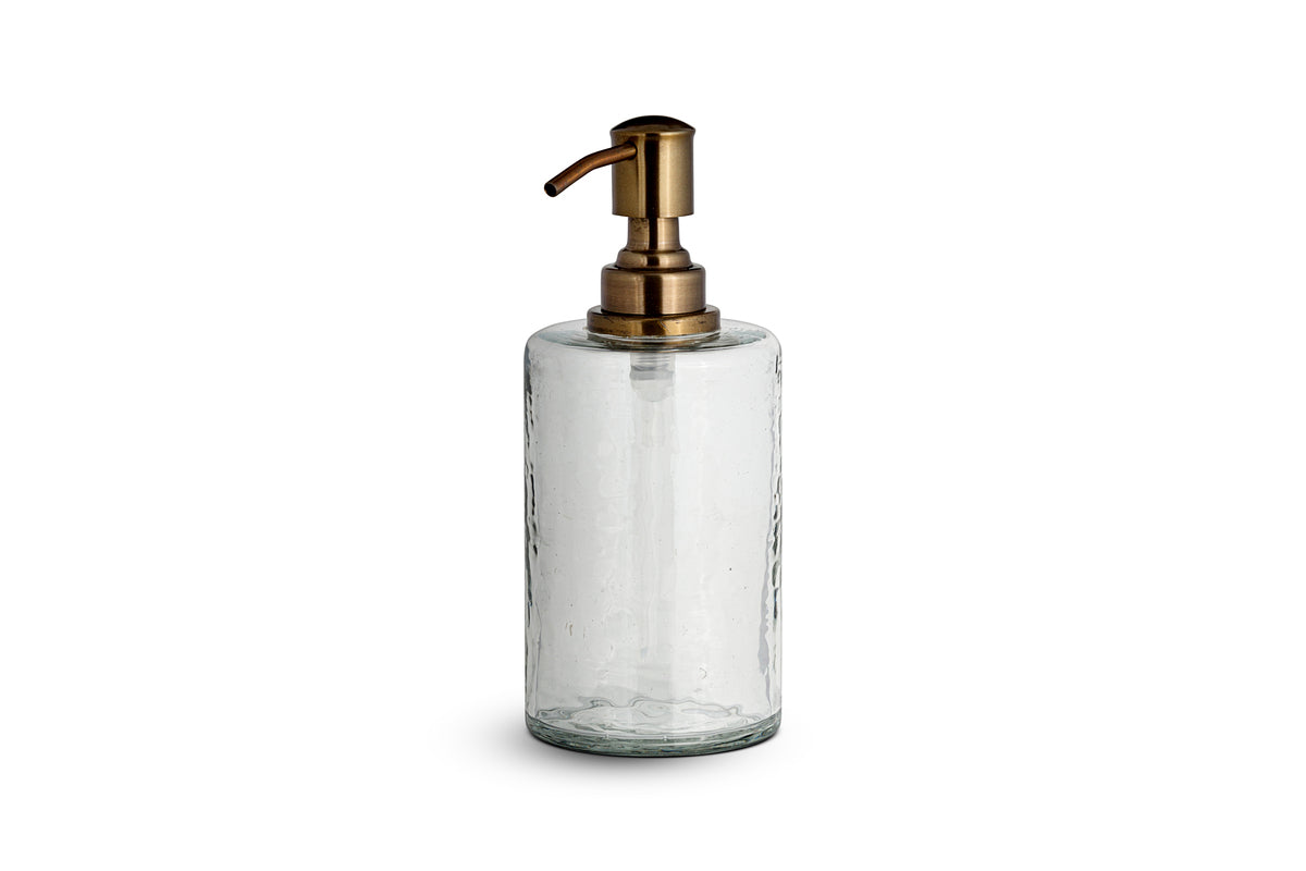 The Ilcoso Recycled Hammered Glass Soap Dispenser by nkuku features clear hammered glass, a cylindrical shape, and an antique brass pump. Its transparent, modern design stands out against a plain white background.