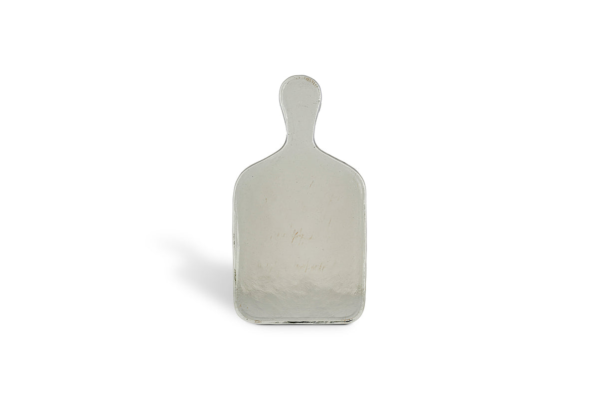 The Komal Serving Platter - Clear by nkuku is a small, rectangular glass platter with a rounded handle, standing upright on a plain white background and casting a faint shadow to the left.