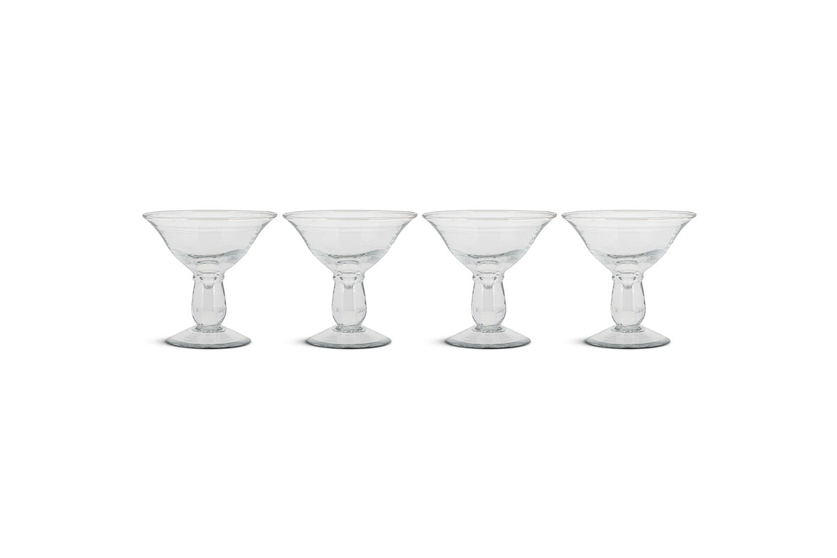 The nkuku Sooraj Champagne Coupe set includes four clear glasses with vintage charm, featuring wide, shallow bowls, short stems, and sturdy round bases. They’re arranged in a row against a plain white background.