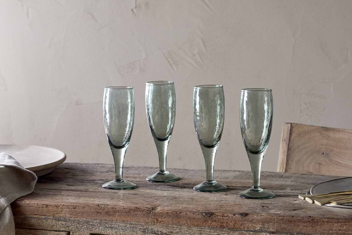 A set of four nkuku Yala Hammered Champagne Glasses in Sage Green is displayed on a rustic wooden table with plates, cutlery, and a napkin nearby, set against a light beige textured wall.
