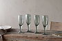 A set of four nkuku Yala Hammered Champagne Glasses in Sage Green is displayed on a rustic wooden table with plates, cutlery, and a napkin nearby, set against a light beige textured wall.