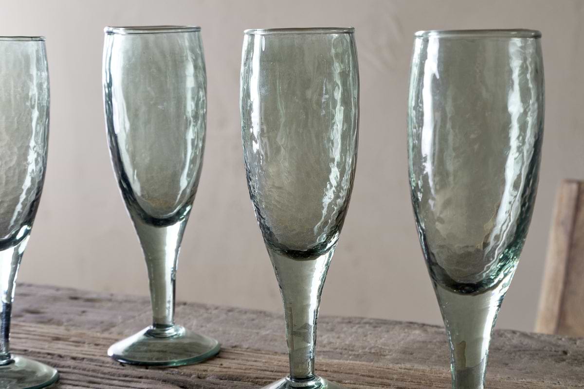 Four Yala Hammered Champagne Glasses in sage green by nkuku stand on a rustic wooden surface against a beige background. The set of four glasses is partially cropped, showing three fully and part of the fourth.