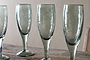 Four Yala Hammered Champagne Glasses in sage green by nkuku stand on a rustic wooden surface against a beige background. The set of four glasses is partially cropped, showing three fully and part of the fourth.