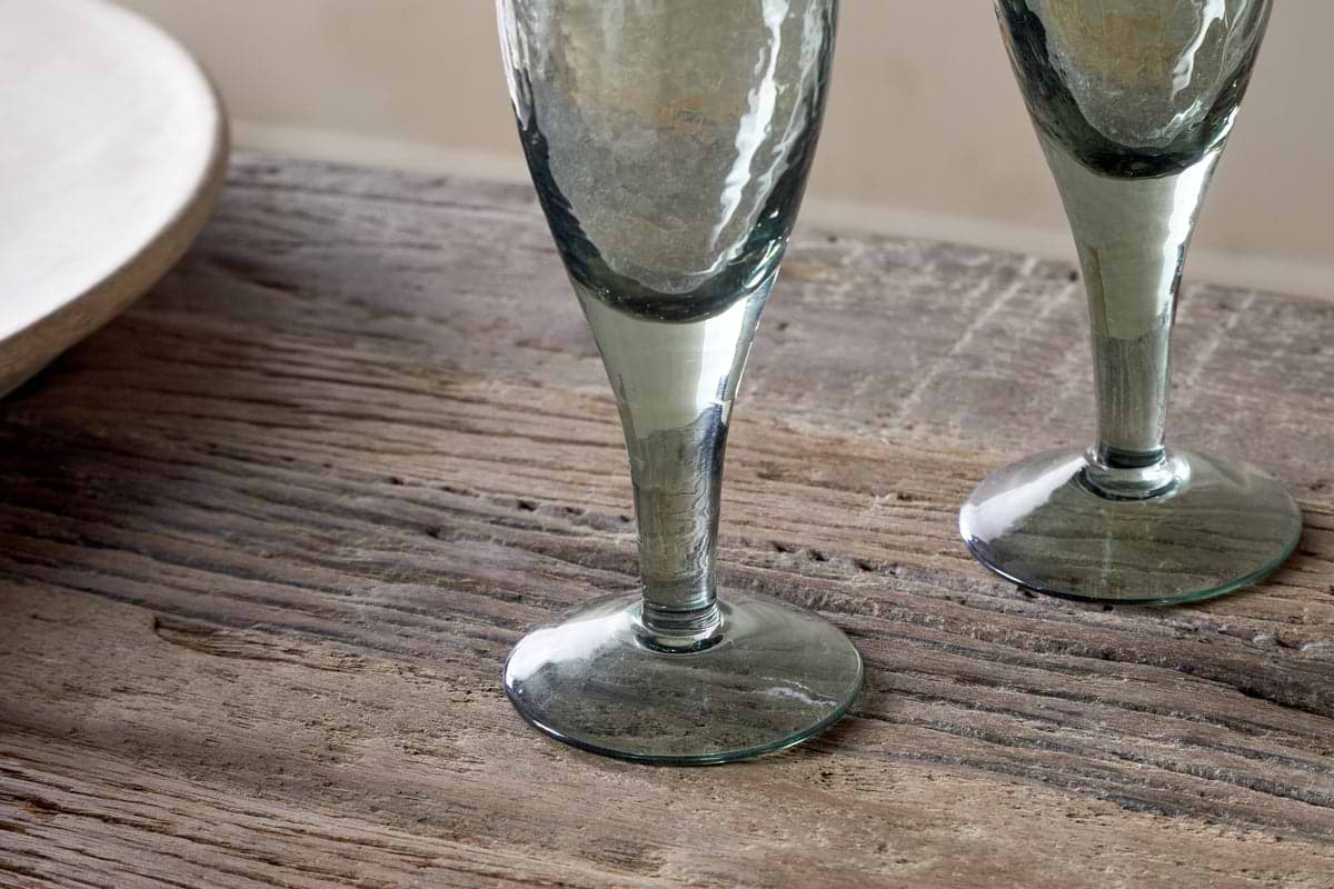 Two Yala Hammered Champagne Glasses in Sage Green by nkuku sit on a rustic wooden table, displaying their recycled glass with subtle hammered detailing. Part of a ceramic plate is visible to the left.