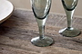 Two Yala Hammered Champagne Glasses in Sage Green by nkuku sit on a rustic wooden table, displaying their recycled glass with subtle hammered detailing. Part of a ceramic plate is visible to the left.