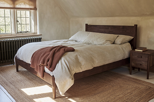 A cozy bedroom features the nkuku Anbu Acacia Bed in Washed Walnut, beige bedding, and a rust blanket at the corner. A wooden nightstand with books sits beside the bed as sunlight filters through window blinds.