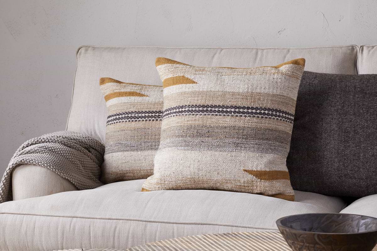 A beige sofa features two nkuku Aravali Wool Cushion Covers in ochre and earthy tones, a gray textured blanket over the arm, a dark gray pillow behind, and a wooden bowl on a light-colored table in front.
