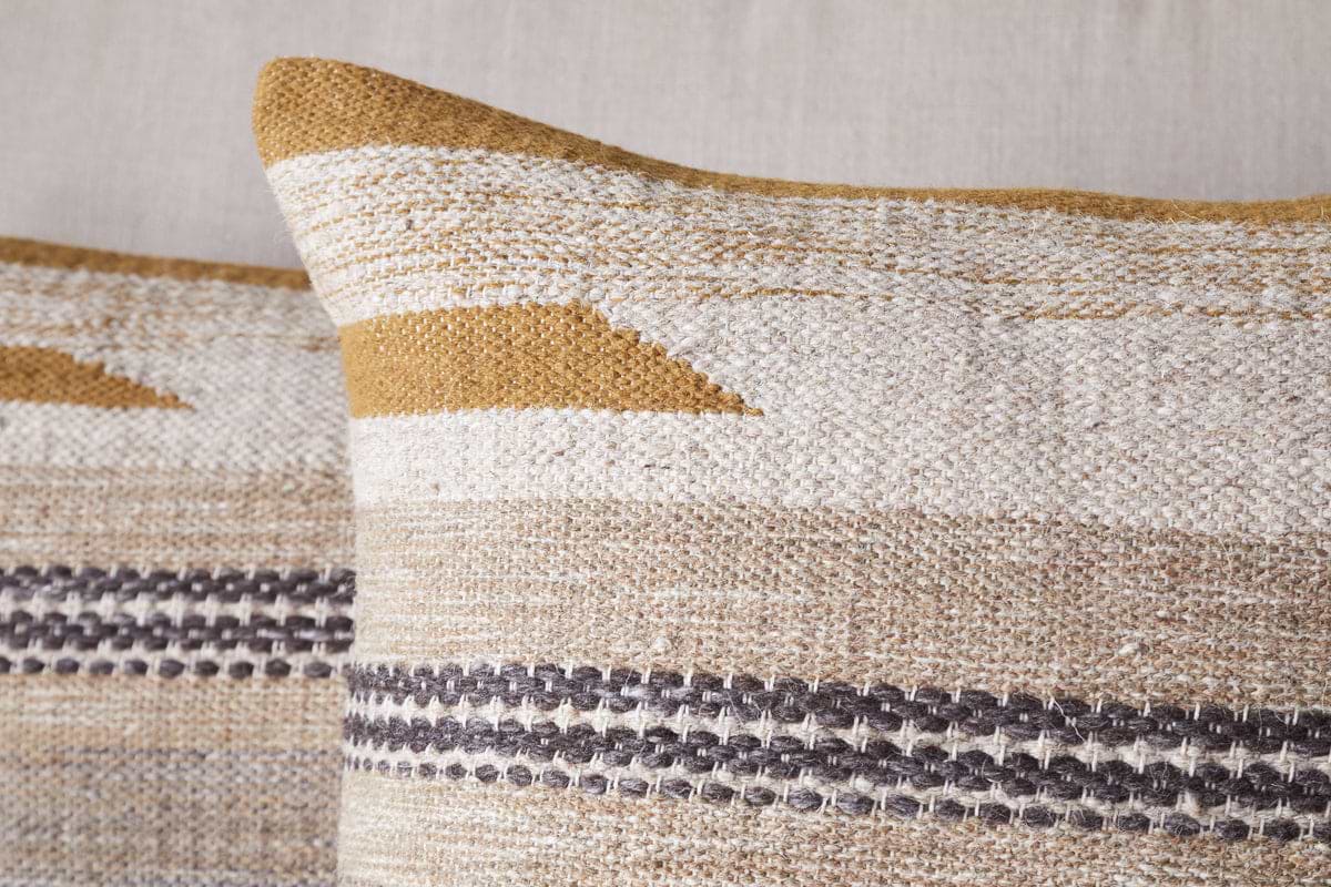 A close-up of two Aravali Wool Cushion Covers in Ochre by nkuku, showing woven geometric designs and horizontal stripes in beige, mustard yellow, and dark brown on a neutral fabric background.