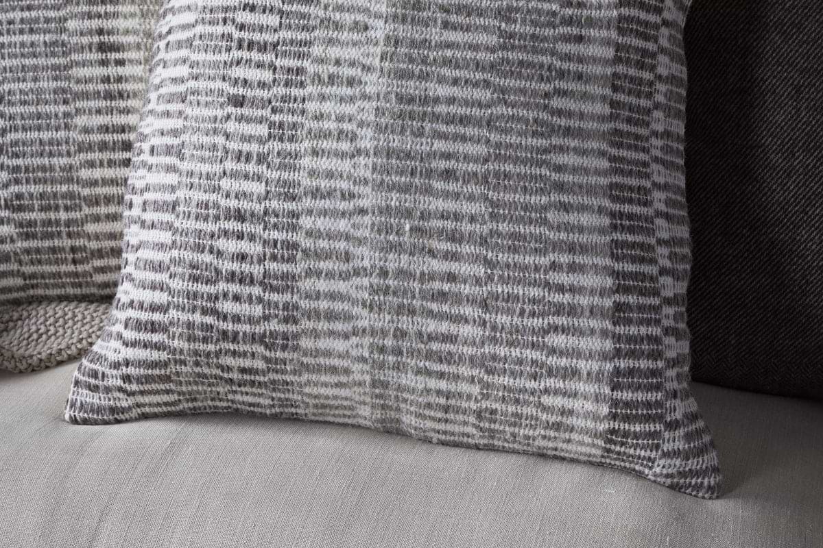 A close-up shows the nkuku Aahna Wool Cushion Cover - Charcoal, featuring a woven gray and white geometric pattern on a light sofa, with another dark pillow behind it. The cover is crafted from wool and recycled polyester.