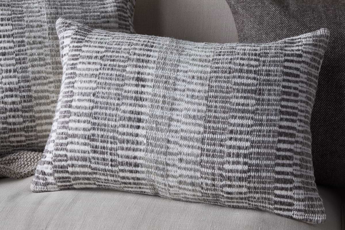 The nkuku Aahna Wool Cushion Cover in Charcoal features a textured, woven design in gray and white wool and recycled polyester, styled here on a light fabric sofa with coordinating cushions in the background.