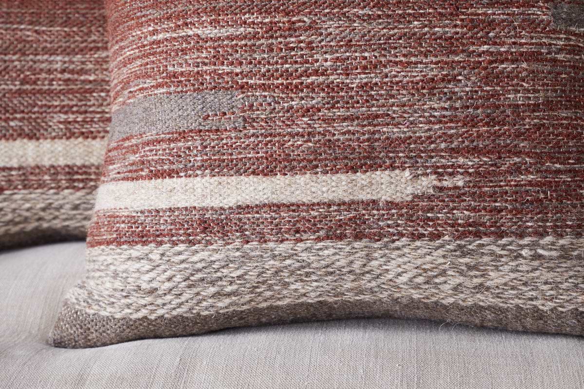 Close-up of the nkuku Aakash Wool Cushion Cover in rust, featuring textured horizontal stripes in earthy brown, beige, and cream tones, shown on a light gray fabric surface.